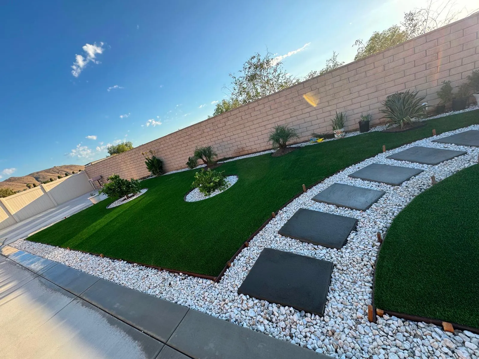 Backyard artificial turf lawn with white gravel border and dark stepping stones along a block wall